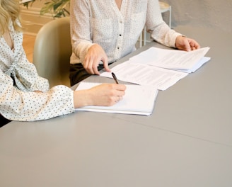 woman signing on white printer paper beside woman about to touch the documents